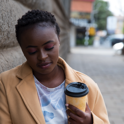 A young women leans against a brick wall with coffee and her phone in hand